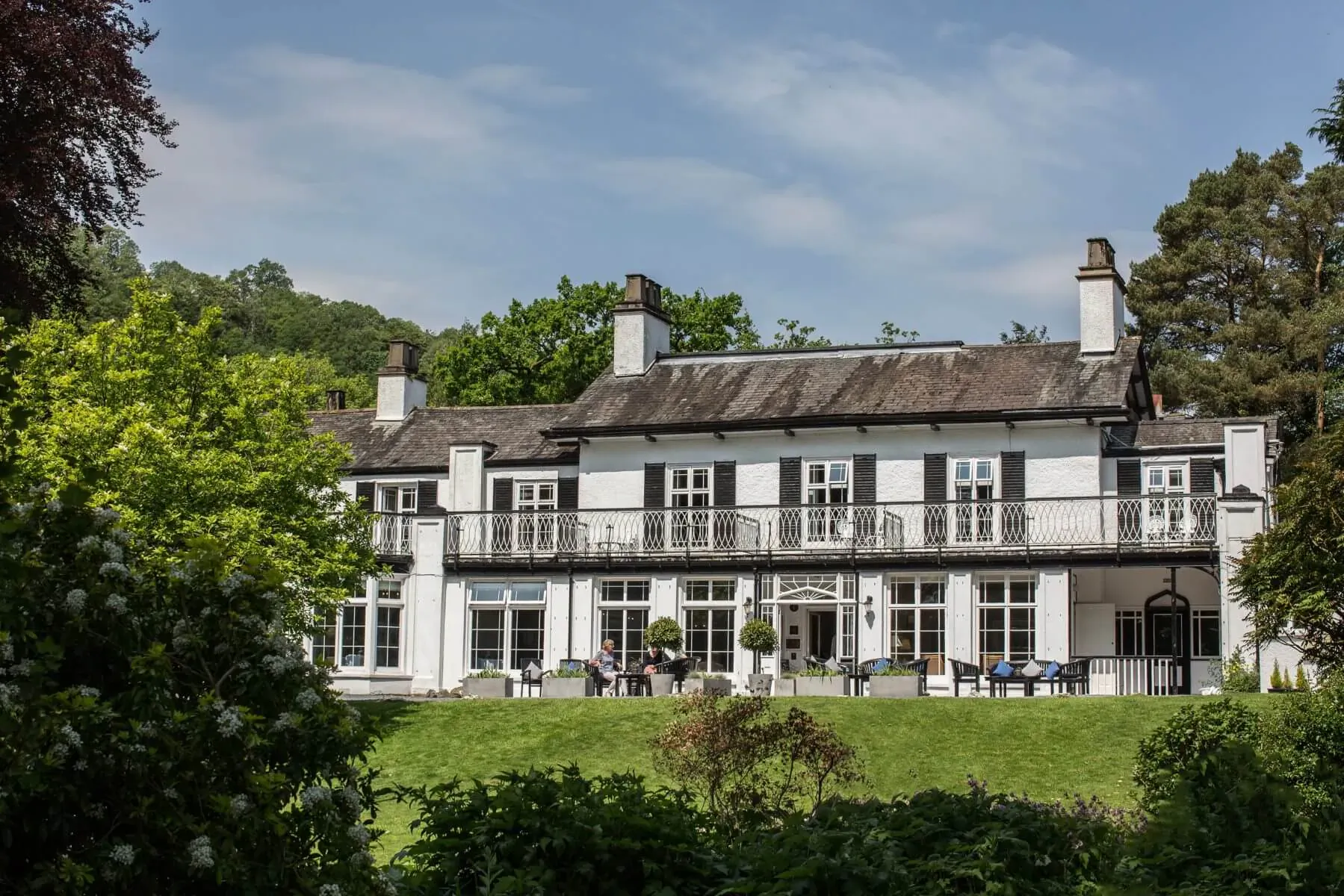 Rothay Manor Boutique Hotel, a white Georgian manor house with balcony, people at outdoor tables, surrounded by green hills and trees.