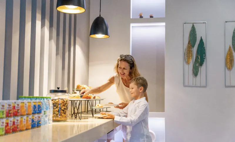 Smiling mother serving breakfast cereals to young boy at modern hotel counter with pendant lights and leaf art.