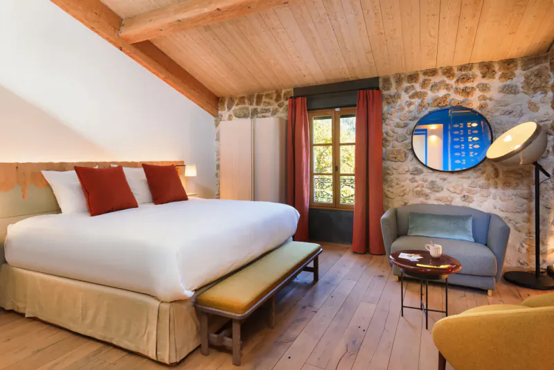 Cozy bedroom at Lou Calen Boutique Hotel in Provence with wooden beams, stone walls, king bed with red pillows, blue mirror, and armchair.