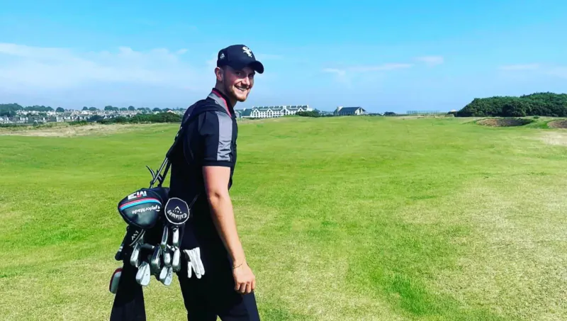 Smiling golfer in black shirt and cap walks on green golf course with golf bag, ocean view backdrop