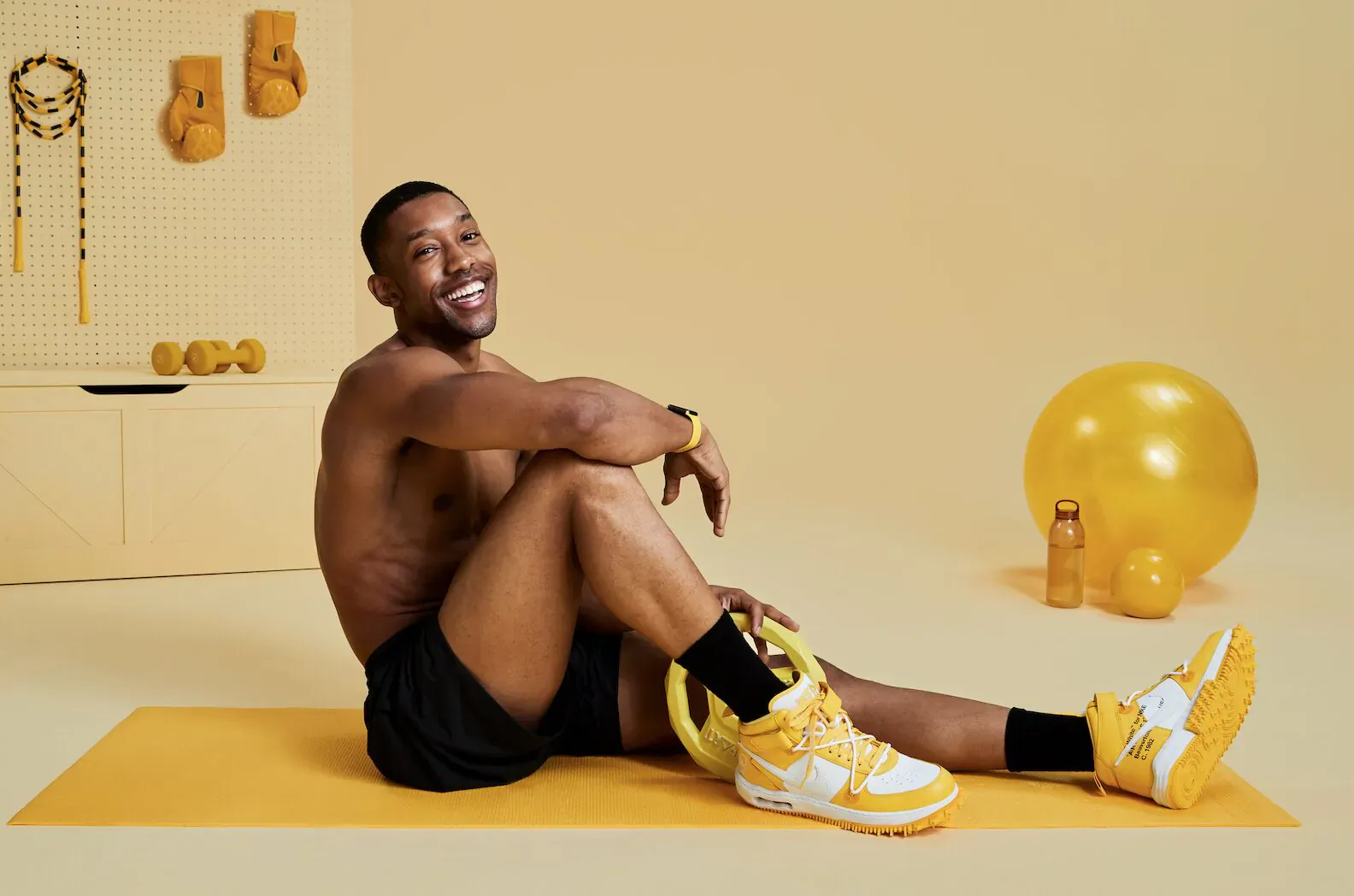 Shirtless Black man smiling, sitting on yellow yoga mat in black shorts and yellow Air Jordan sneakers amid yellow gym props and exercise ball