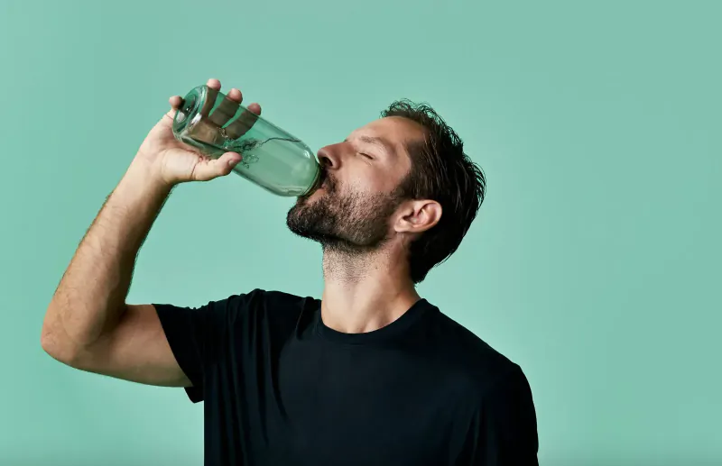Bearded man in black t-shirt drinks from green glass bottle against teal background
