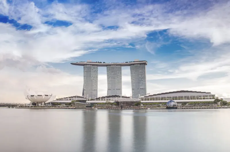 Marina Bay Sands' three iconic towers and infinity pool atop, reflected in calm Marina Bay waters under blue sky with clouds