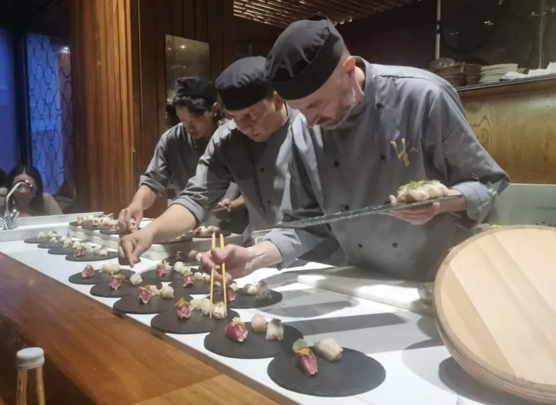Chefs in black hats and grey uniforms preparing sushi and nigiri on black plates at a wooden kitchen counter