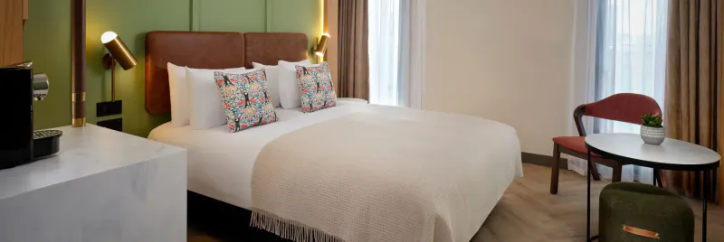 Elegant hotel room with king bed, green headboard, brass lamps, white counter, red armchair, and plant by window.