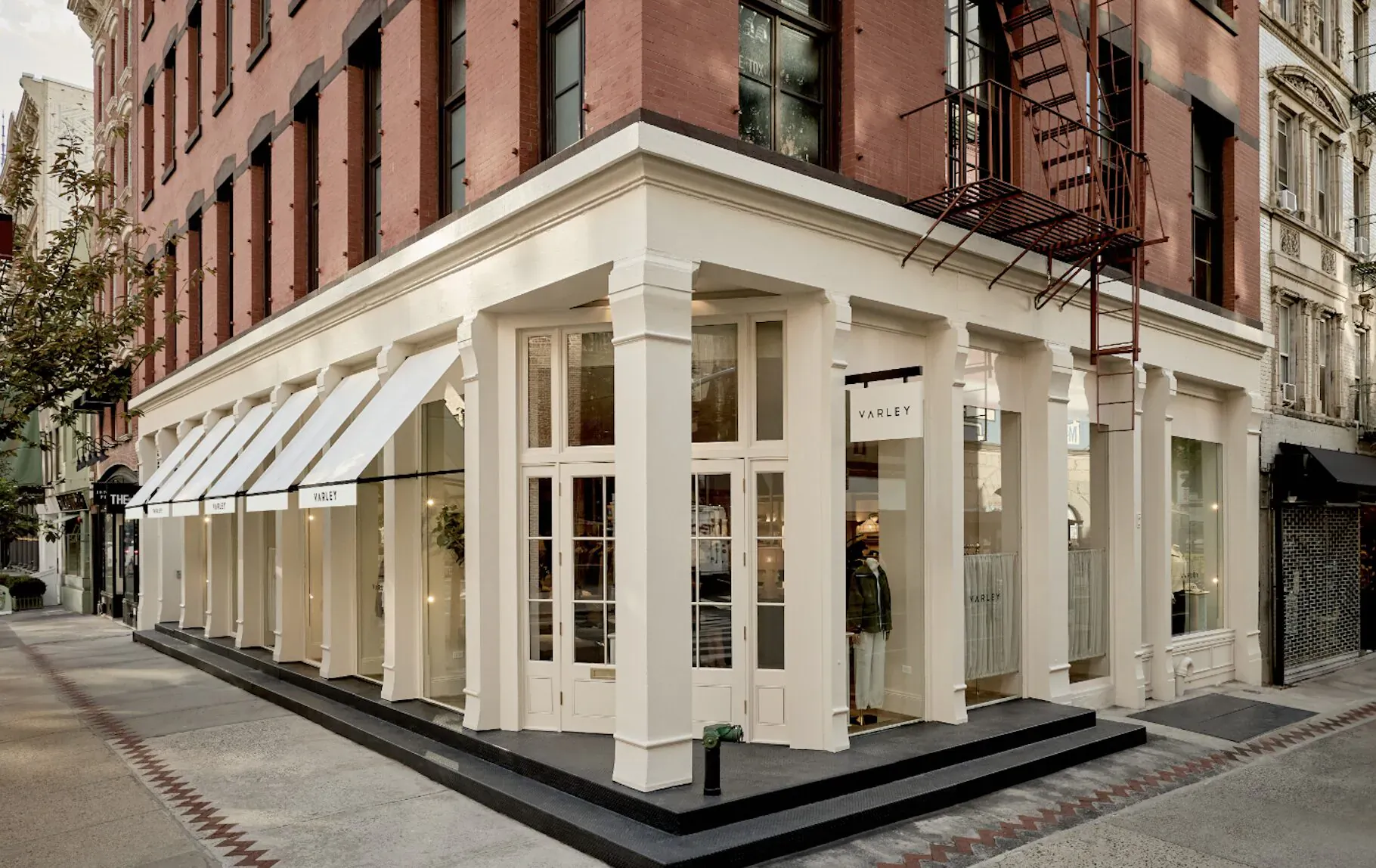 Varley flagship store exterior in SoHo, white corner building with awnings, large windows displaying clothes, red brick walls, fire escape.