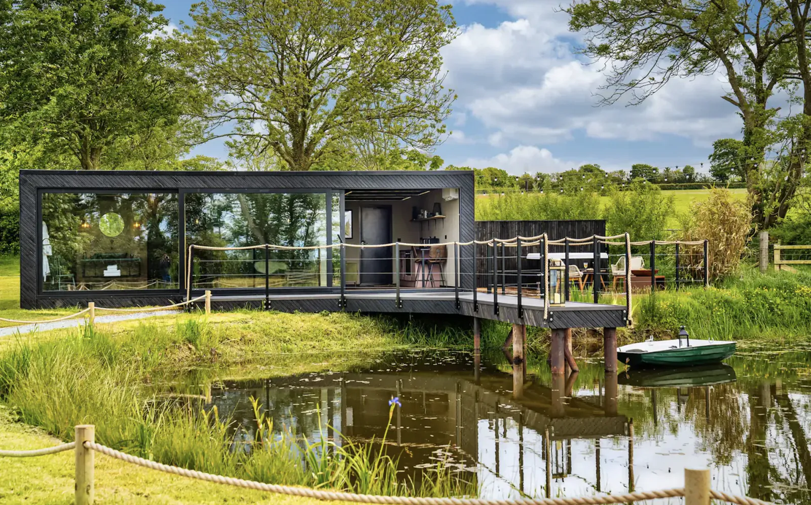 Modern black luxury glamping pod on wooden deck over pond, surrounded by green trees and fields under partly cloudy sky.