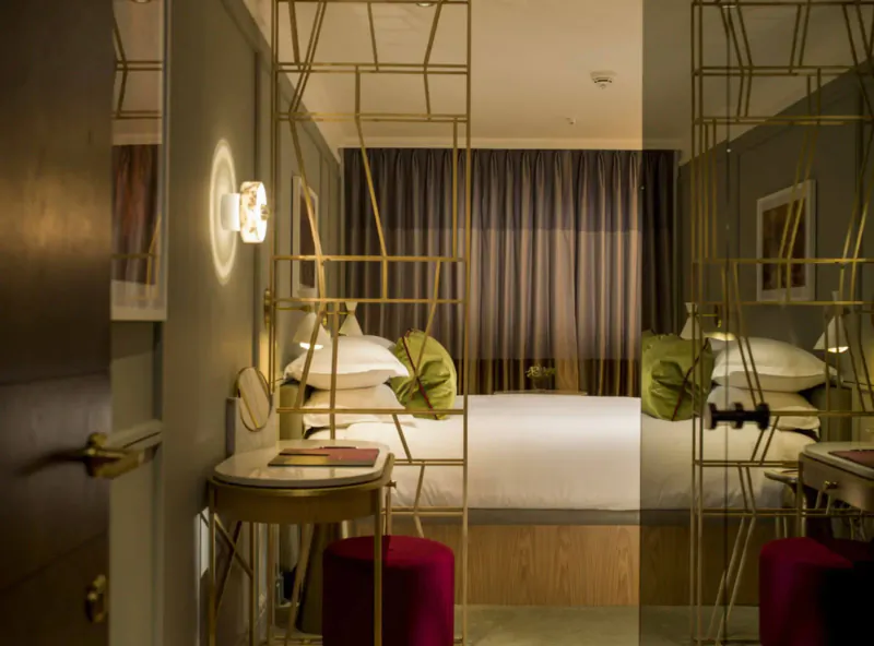 Elegant hotel room at The Guardsman with gold-framed mirrors, white bed, green pillows, red stools, and warm lighting.