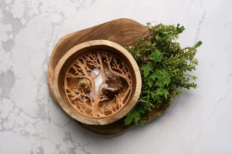 Wooden bowl with tree-like cutout design holding creamy soup garnished with fresh green herbs on a wooden board atop white marble.