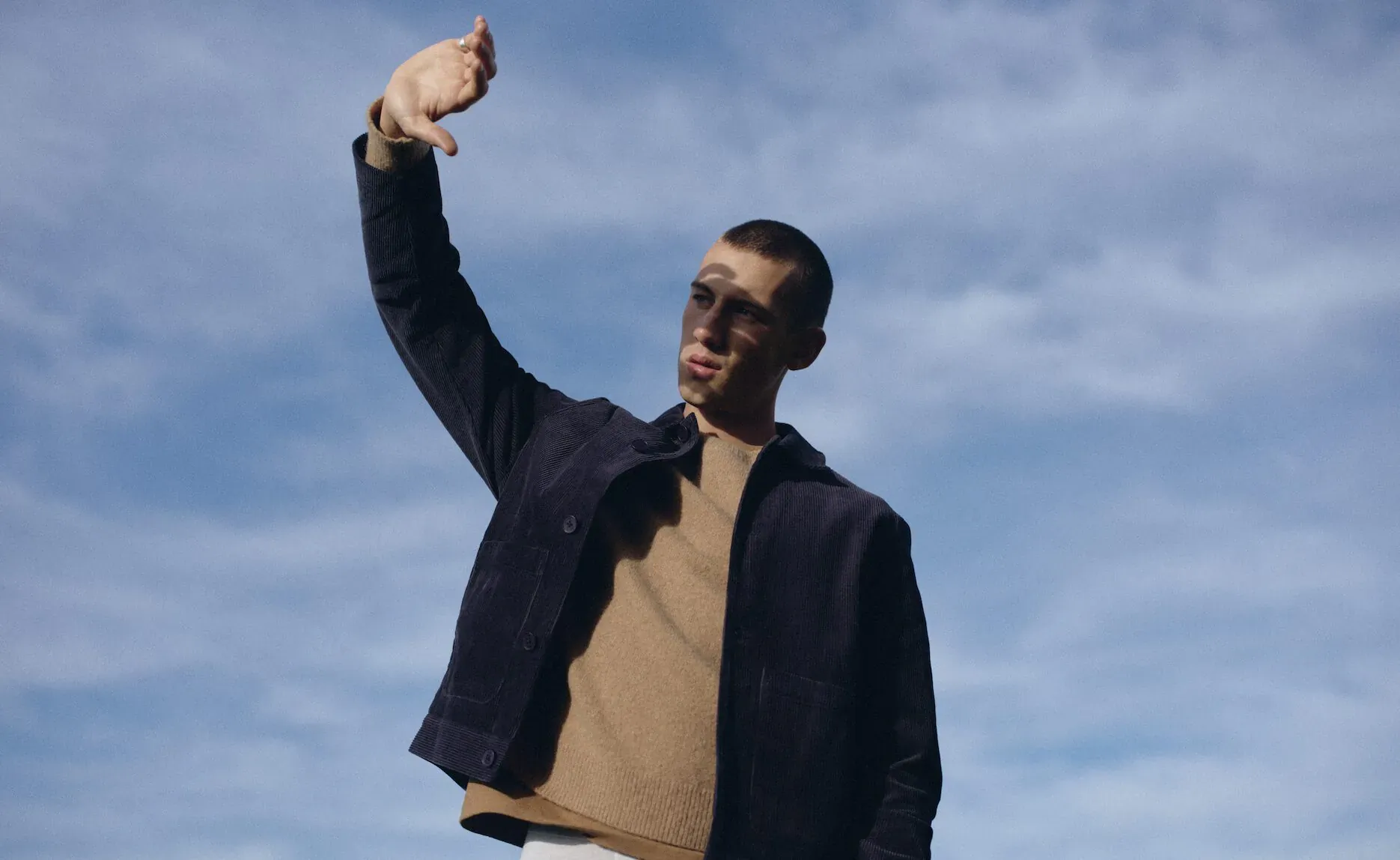 Man in navy Wax London jacket and beige shirt raises arm against cloudy sky in Canary Wharf