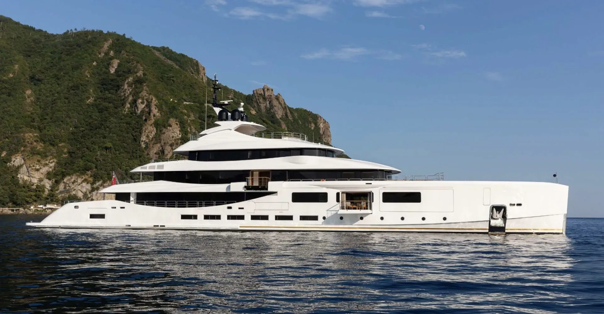 White luxury superyacht ALFA docked in calm waters by lush green cliffs under blue sky.
