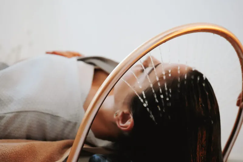 Woman lying back in salon chair during scalp treatment with acupuncture needles in hair, framed by copper hoop