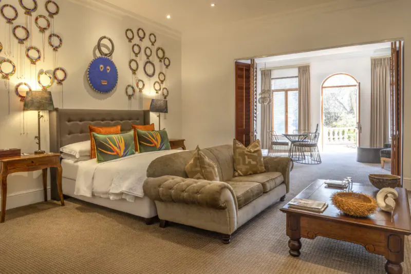 Elegant bedroom with white bed, orange pillows, beige sofa, wooden table, wall art, and balcony view.
