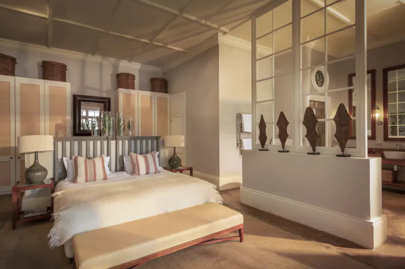 Elegant bedroom in Samsara Karoo Safari Villa with white king bed, striped pillows, wooden sculptures, and large windows.
