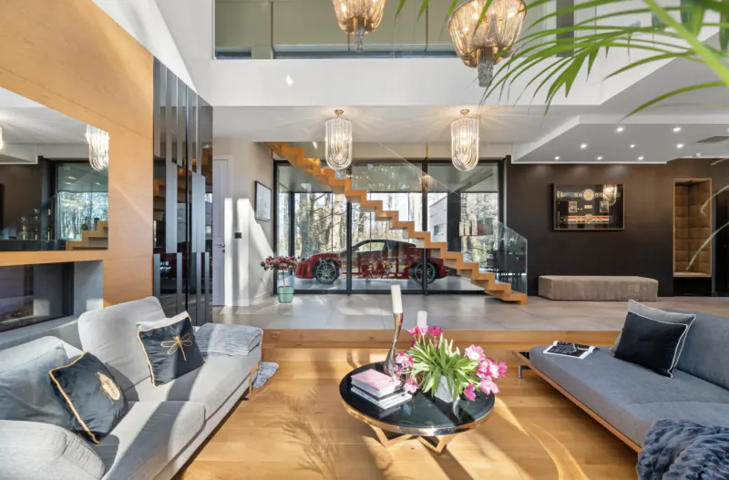 Modern open-plan living room with wooden floors, white sofas, flowers on table, red sports car visible through glass staircase.