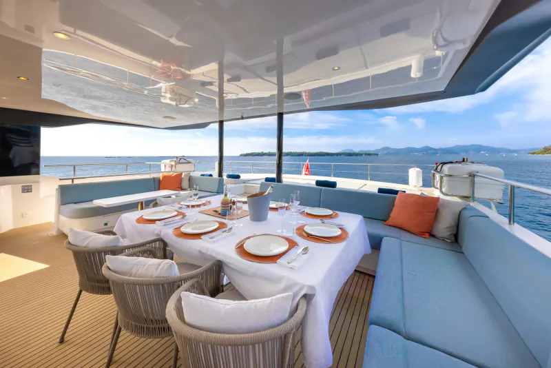 Elegant yacht deck dining table set with white plates, orange accents, blue cushions, and sea-mountain view.