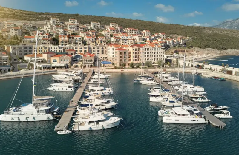 Aerial view of a marina with white yachts docked in calm waters, surrounded by terraced red-roofed buildings on a hillside.