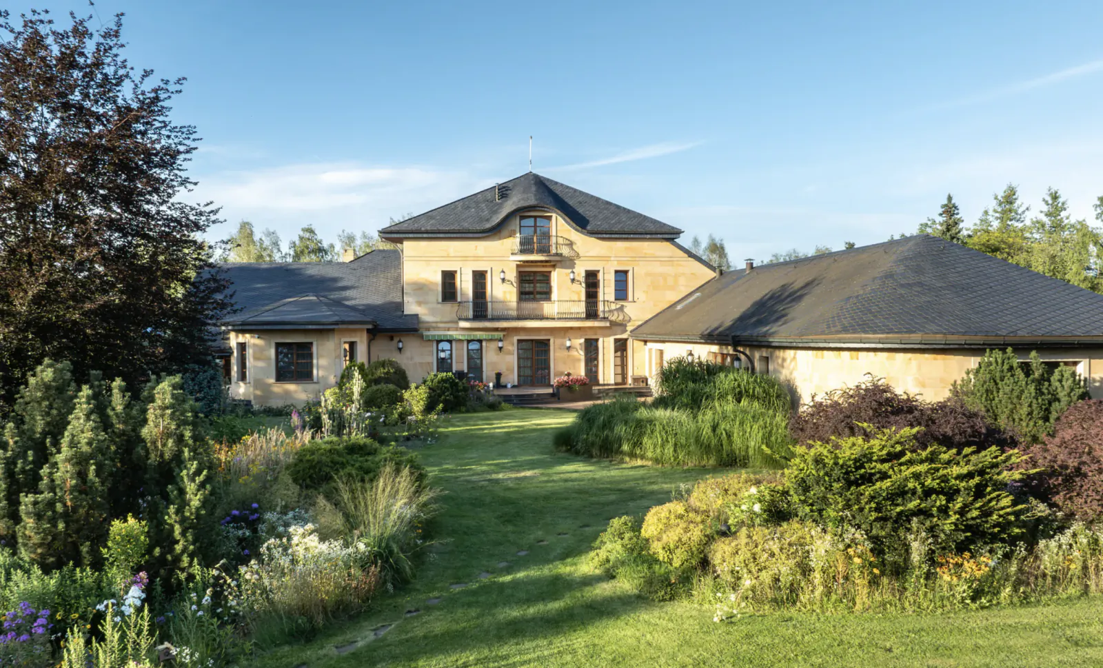 Beige luxury house with dark roofs, balcony, and landscaped garden under blue sky with trees.