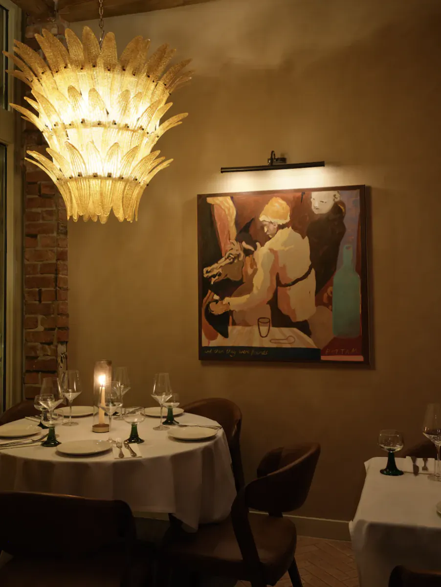 Elegant dining room at Ottega 35 with large feather chandelier, abstract painting of man and horse, candlelit white table, wine glasses.