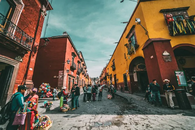 Vibrant cobblestone street in Mexico lined with red and yellow colonial buildings, people and colorful vendors.