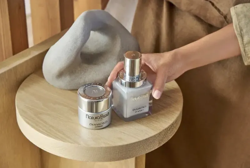 Woman's hand holding Touch Beauty cream jar next to another on wooden table with gray sculpture.