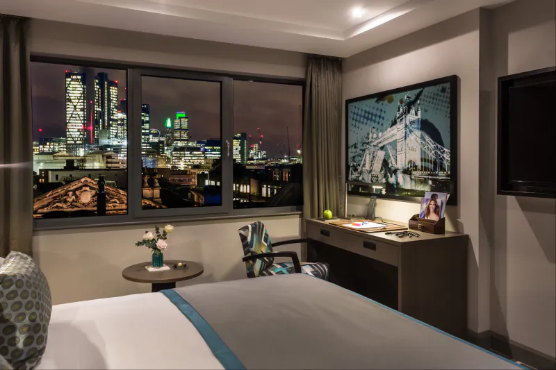 Courthouse Shoreditch hotel room with panoramic nighttime London skyline view from window, bed, desk, and art.