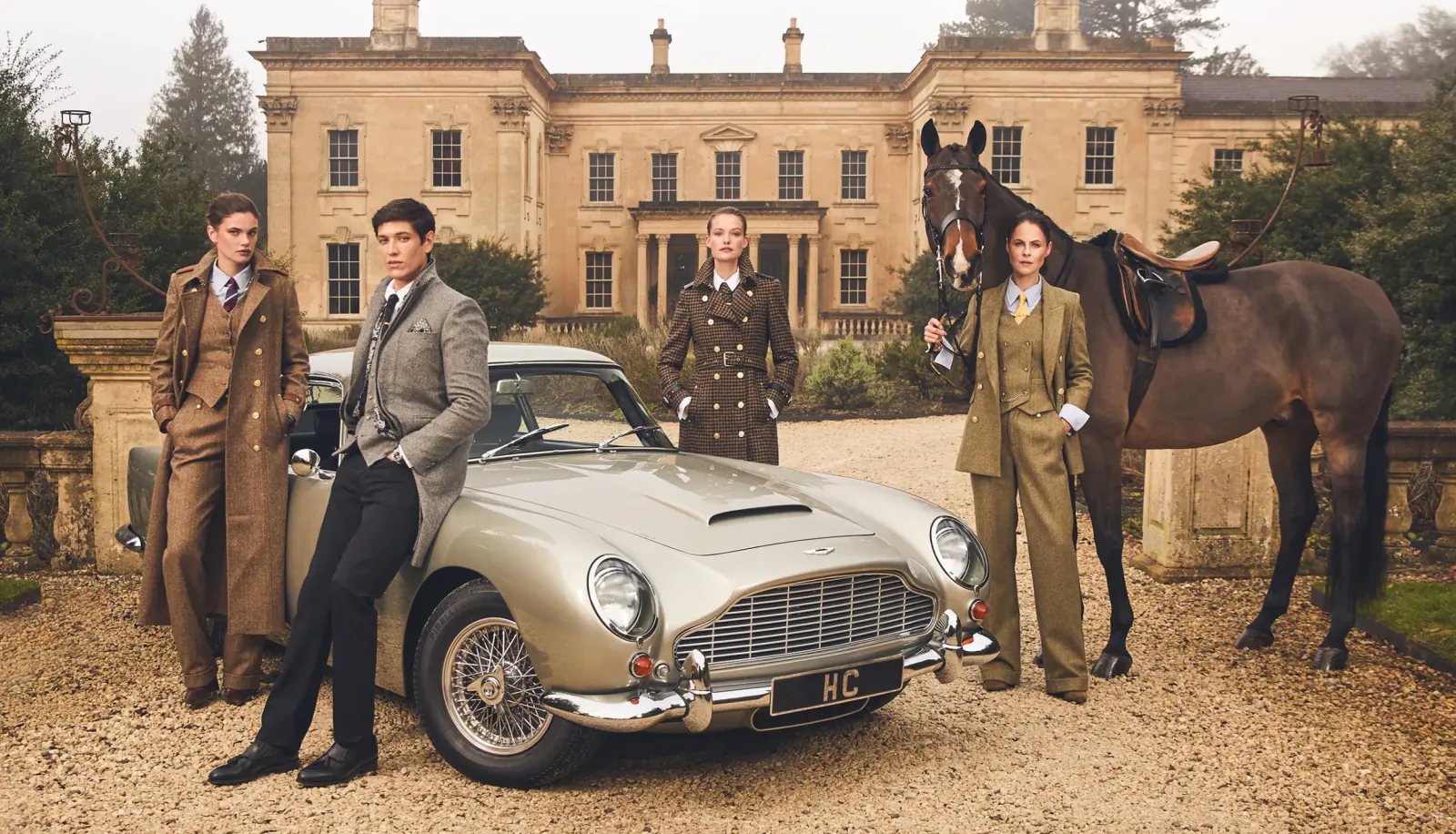 Four stylish men in tweed suits pose by silver Aston Martin and saddled horse in front of stately manor house.