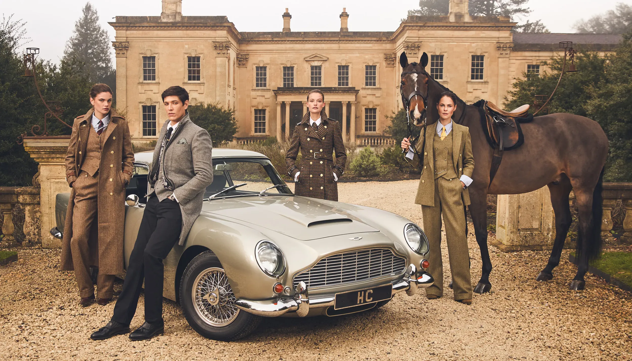 Four stylish men in tweed suits pose by silver Aston Martin and saddled horse in front of stately manor house.