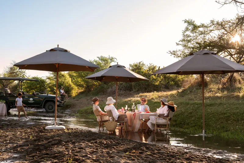 Group dining at elegant outdoor table under umbrellas by river at Samsara Karoo Safari Villa, safari jeep nearby