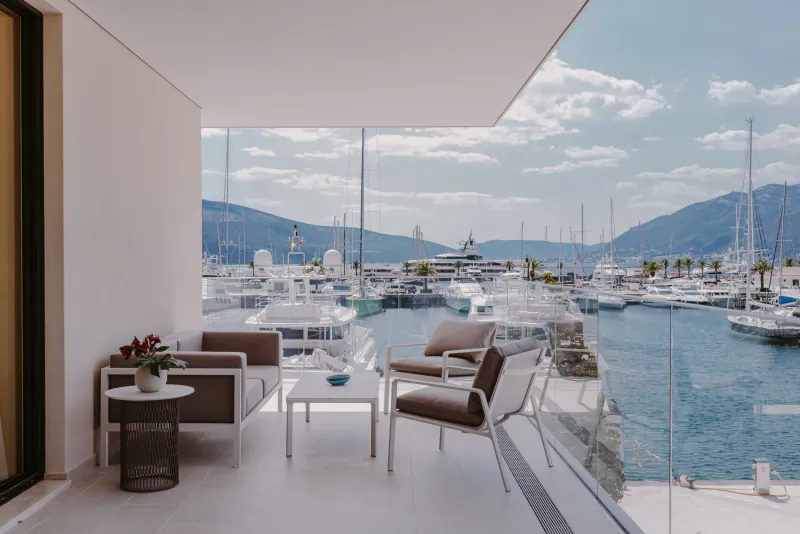 Modern balcony with brown lounge chairs, white table, flowers overlooking marina with yachts and mountains under blue sky.
