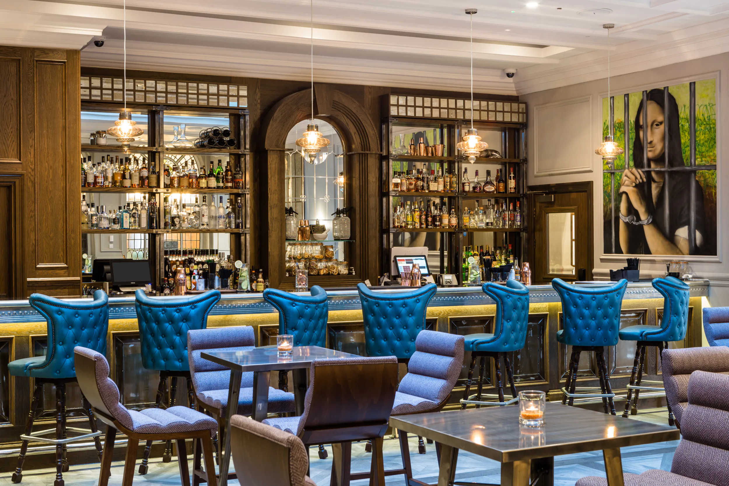 Elegant Courthouse Shoreditch bar with wooden shelves of bottles, blue upholstered bar stools, tables with candles, and large portrait wall mural.