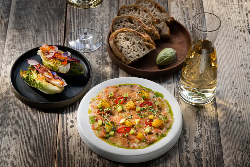Close-up of tuna tartare with veggies on white plate, lettuce wraps on black plates, bread, and white wine on wooden table