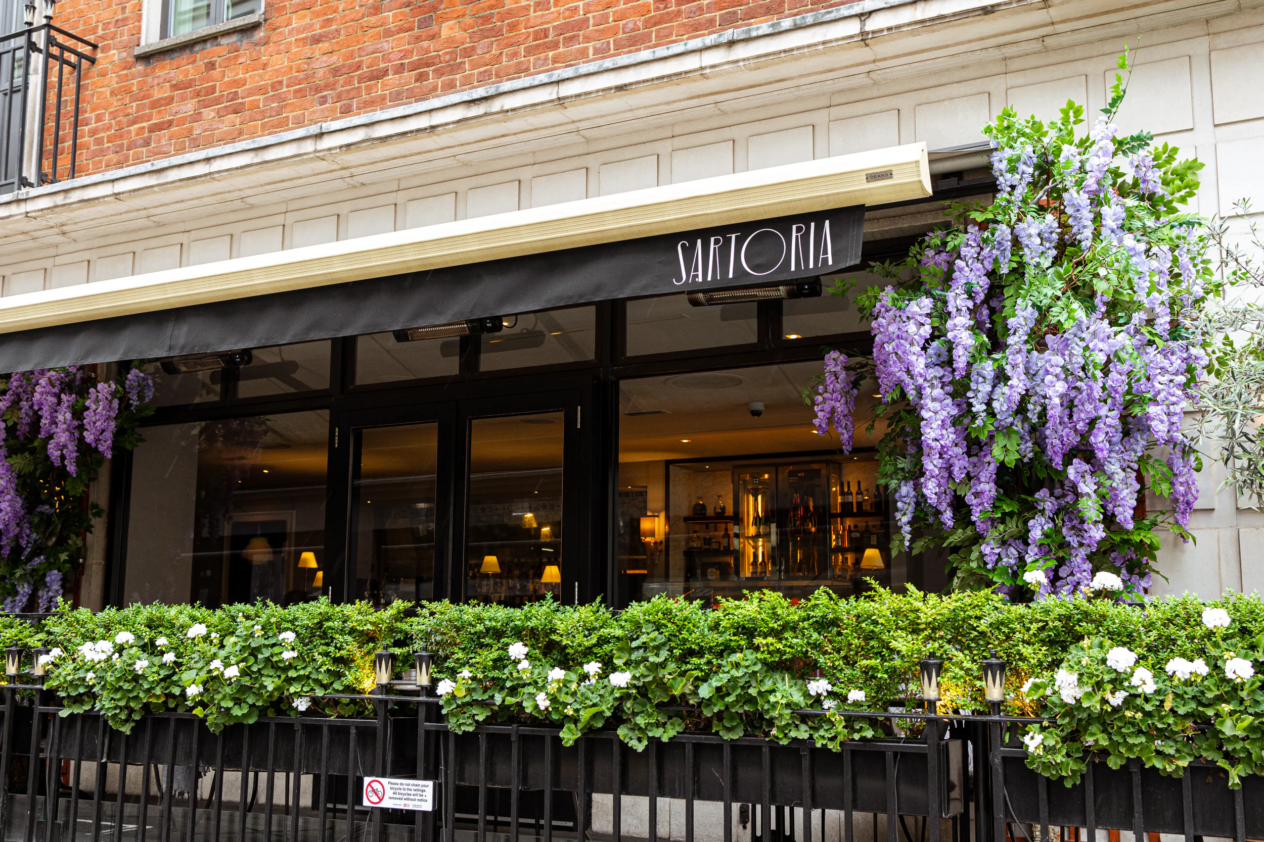 Sartoria restaurant facade on Savile Row, London: black awning, purple wisteria, white flowers in railing, elegant brick building.