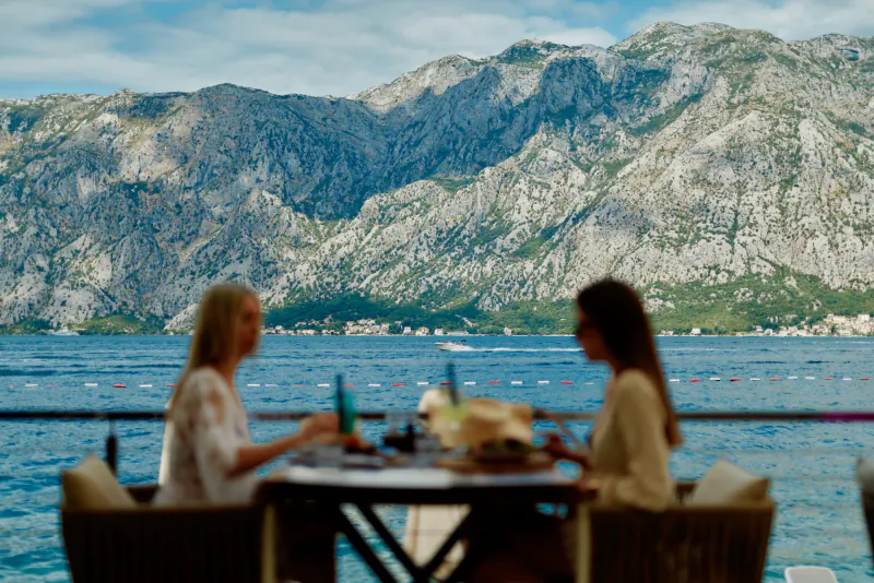 Two women in summer dresses dine at outdoor table overlooking Kotor Bay, Montenegro mountains and sea.