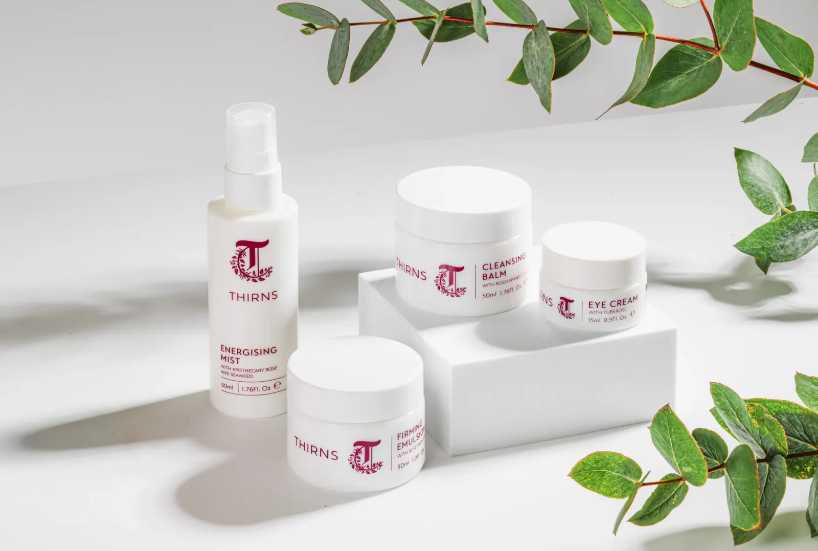 Thirns skincare products: spray bottle, cream jars on white stand with eucalyptus leaves.