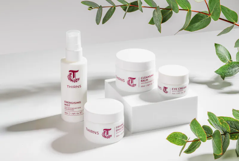 Thirns skincare products: spray bottle, cream jars on white stand with eucalyptus leaves.