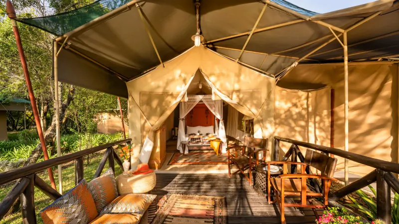 Luxurious beige safari tent at Fairmont Mara Safari Club, Kenya, with deck featuring chairs, pillows, and open bedroom, surrounded by greenery.
