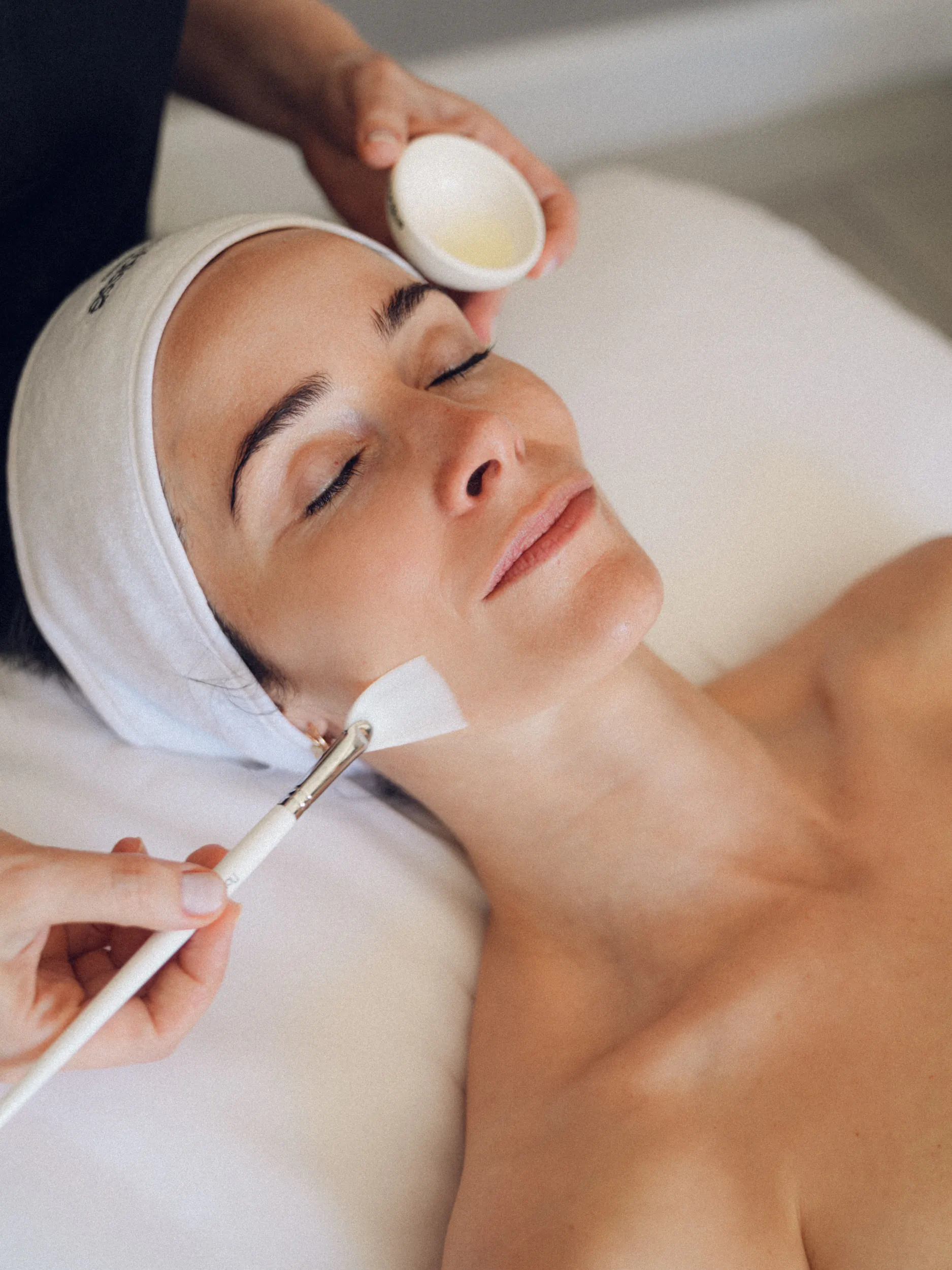 Esthetician applies facial mask with brush to relaxed woman wearing headband on spa bed