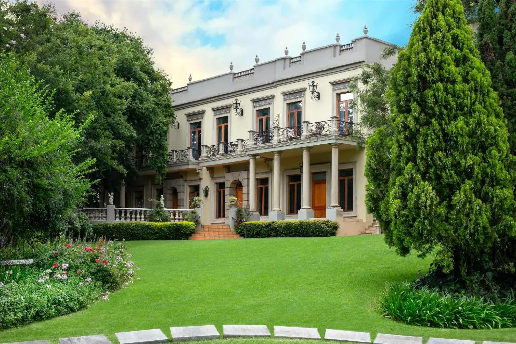 Elegant beige mansion with balconies and columns, surrounded by lush green lawns, trees, and flower beds under a blue sky.