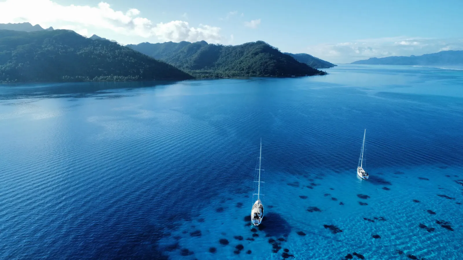 Aerial view of two sailboats anchored in turquoise lagoon with lush green mountains and clear blue waters.