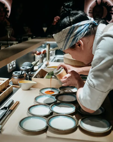 Chef in white uniform and blue headband pours sauce from small jug onto multiple ceramic plates in modern kitchen.