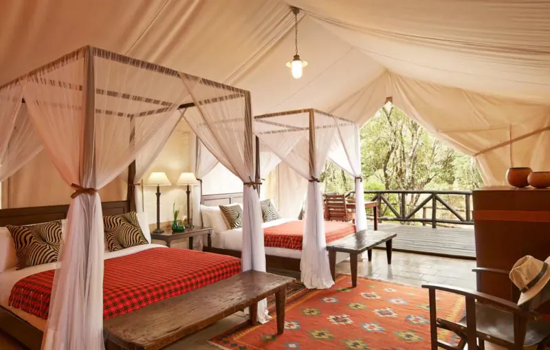 Luxurious tent interior at Fairmont Mara Safari Club with two four-poster beds, wooden furniture, and deck view of Kenyan wilderness.