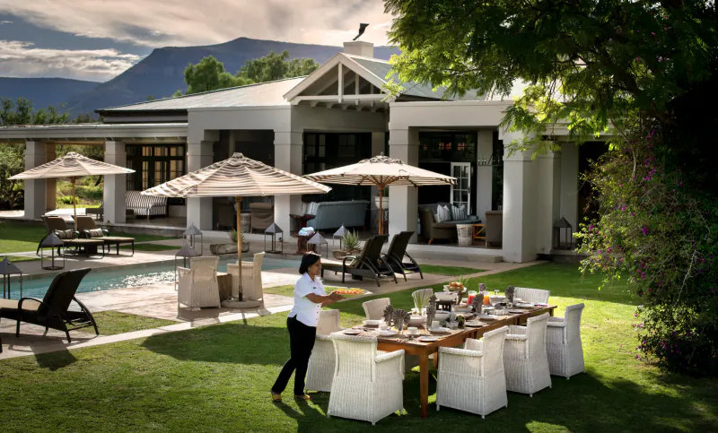 Server in white uniform sets dining table outdoors at Samsara Karoo Safari Villa, pool, loungers, mountains beyond.