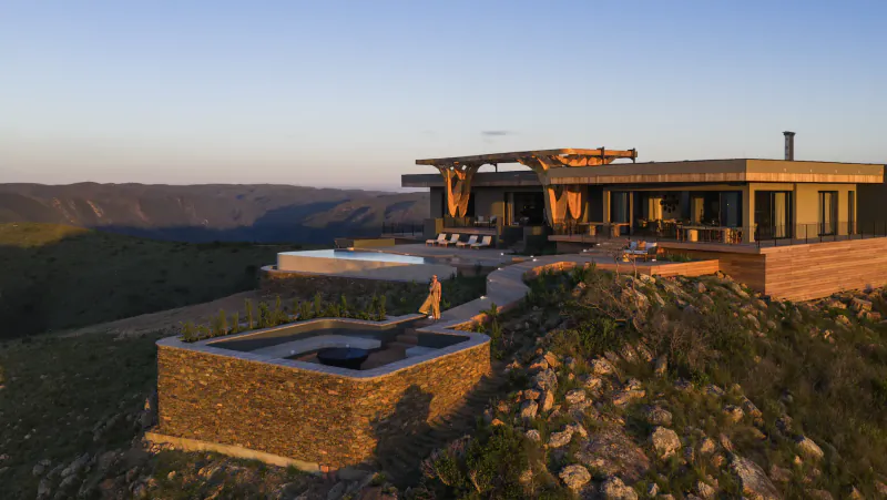 Modern wooden house with infinity pool and fire pit on grassy hillside overlooking vast valley at dusk, Hiddn in Addo