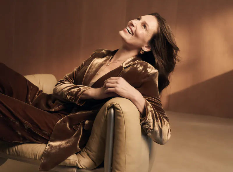 Smiling woman in brown velvet robe reclining on beige sofa, warm lighting, skincare ad vibe