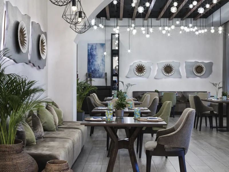 Elegant dining area at The Chedi Luštica Bay with wooden tables, green chairs, sofa, plants, sunburst wall art, and pendant lights.