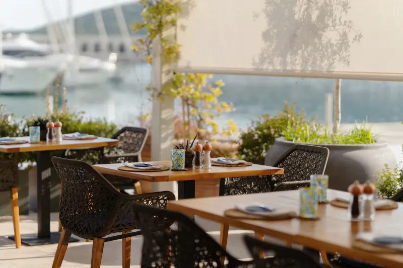 Outdoor dining at The Chedi Luštica Bay Montenegro: wicker chairs, wooden tables with plates, overlooking marina and yachts.