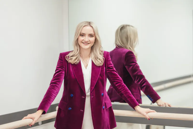 Blonde woman in purple velvet blazer smiling, mirrored reflection, standing by ballet barre in studio