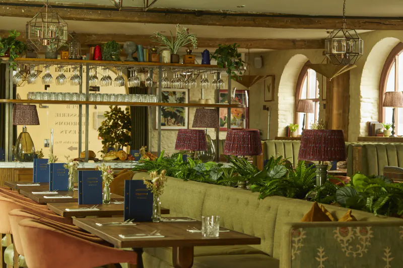 Cozy Kings Head Cirencester inn dining area with wooden beams, potted plants, lamps, and blue menus on tables.