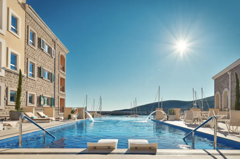 Infinity pool at luxury seaside resort with sun loungers, yachts in marina, stone buildings, and sunny blue sky.
